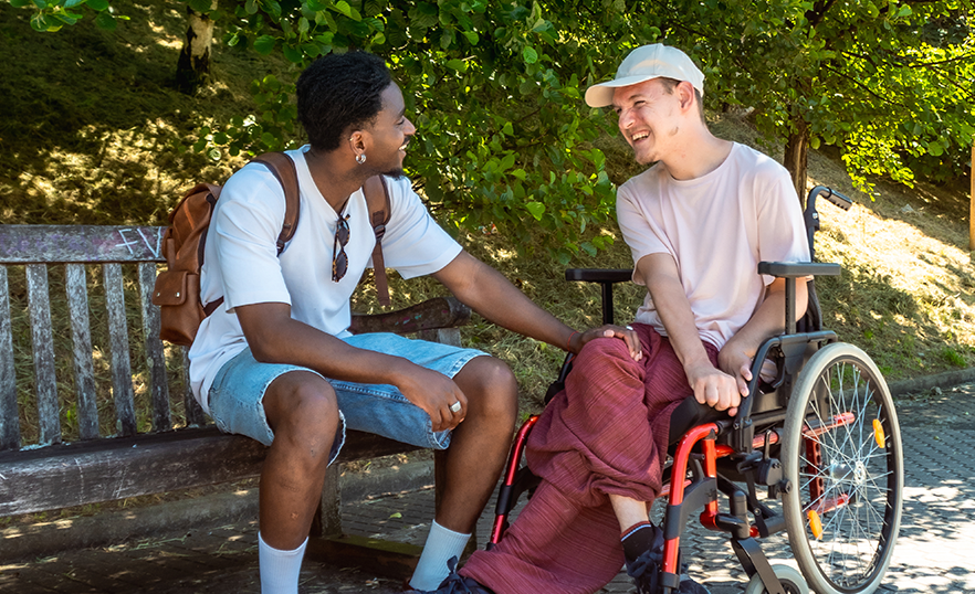 A person on a wheelchair is chatting with his caregiver at a park