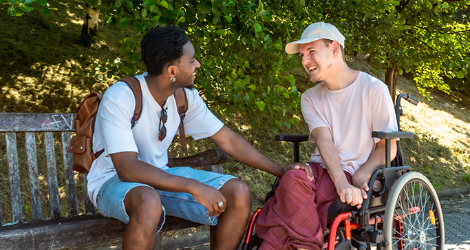 A person on a wheelchair is chatting with his caregiver at a park